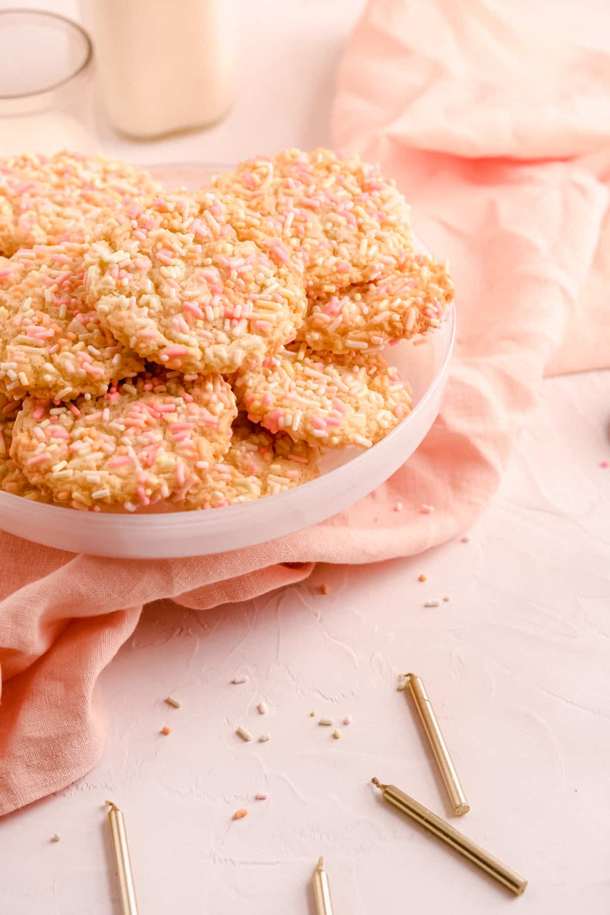 a cake plate of birthday cake cookies covered in rainbow sprinkles with candles in the foreground and glasses of milk in the background