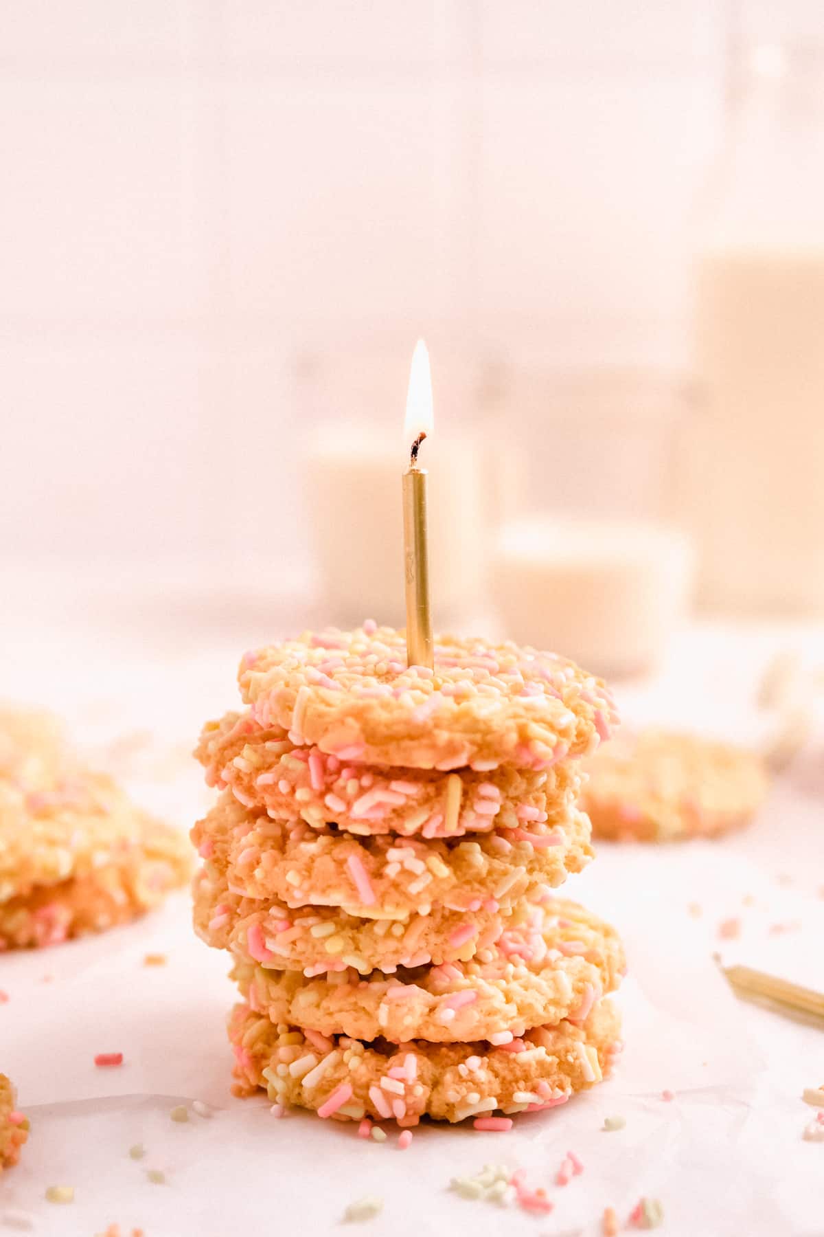 a stack of birthday cake sugar cookies on parchment covered in rainbow sprinkles with a lit birthday candle stuck in it with glasses of milk in the background