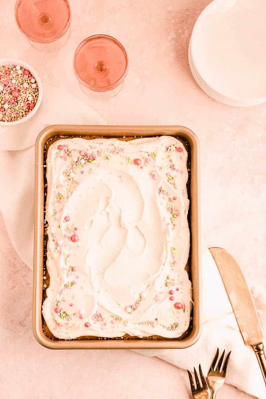 easy vanilla sheet cake with vanilla buttercream and sprinkles on top in a 9" x 13" pan with glasses of rose and a bowl of sprinkles