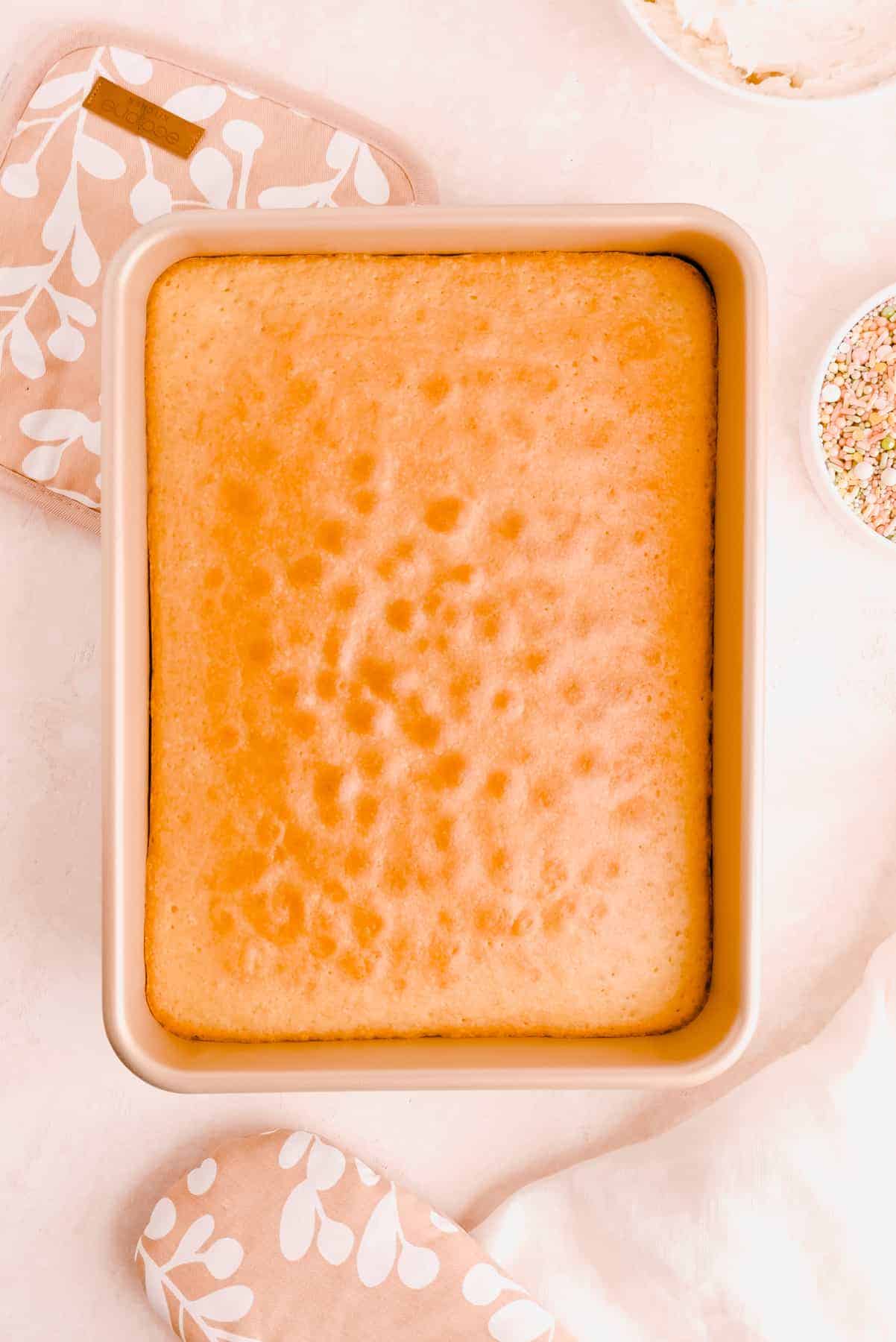 easy vanilla sheet cake in a 9" x 13" pan with an oven mitt, bowl of vanilla frosting and bowl of sprinkles