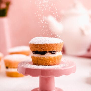 mini victoria sponge cakes filled with jam from above with powdered sugar falling on top of a mini cake plate with a teapot in the background
