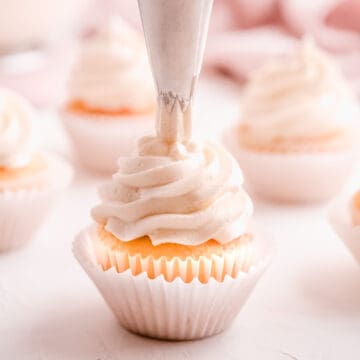 easy vanilla buttercream frosting being swirled on cupcakes from a pastry bag fitted with a star tip with other frosted cupcakes in the background