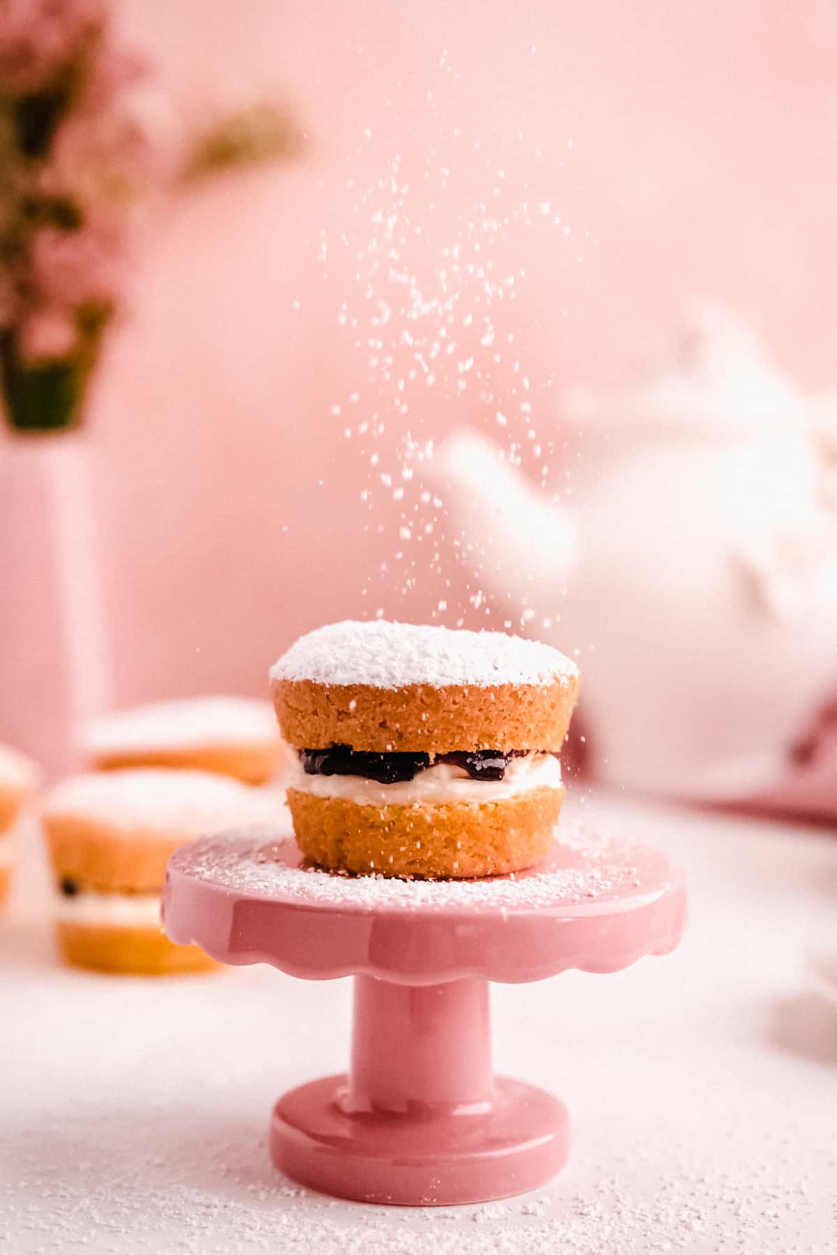 mini victoria sponge cakes filled with jam from above with powdered sugar falling on top of a mini cake plate with a teapot in the background