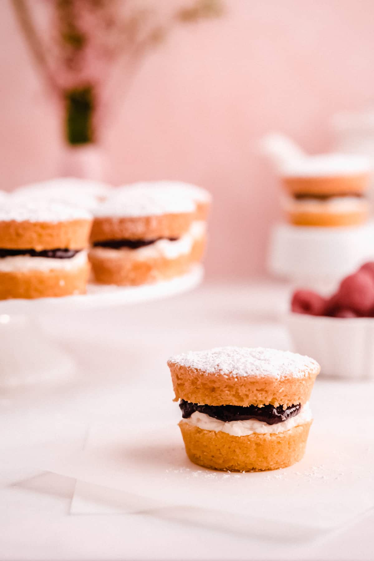 mini victoria sponge cakes filled with jam on a table and cake plate with raspberries and a teapot in the background