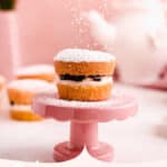 mini victoria sponge cakes filled with jam with powdered sugar falling on top of a mini cake plate with a teapot in the background