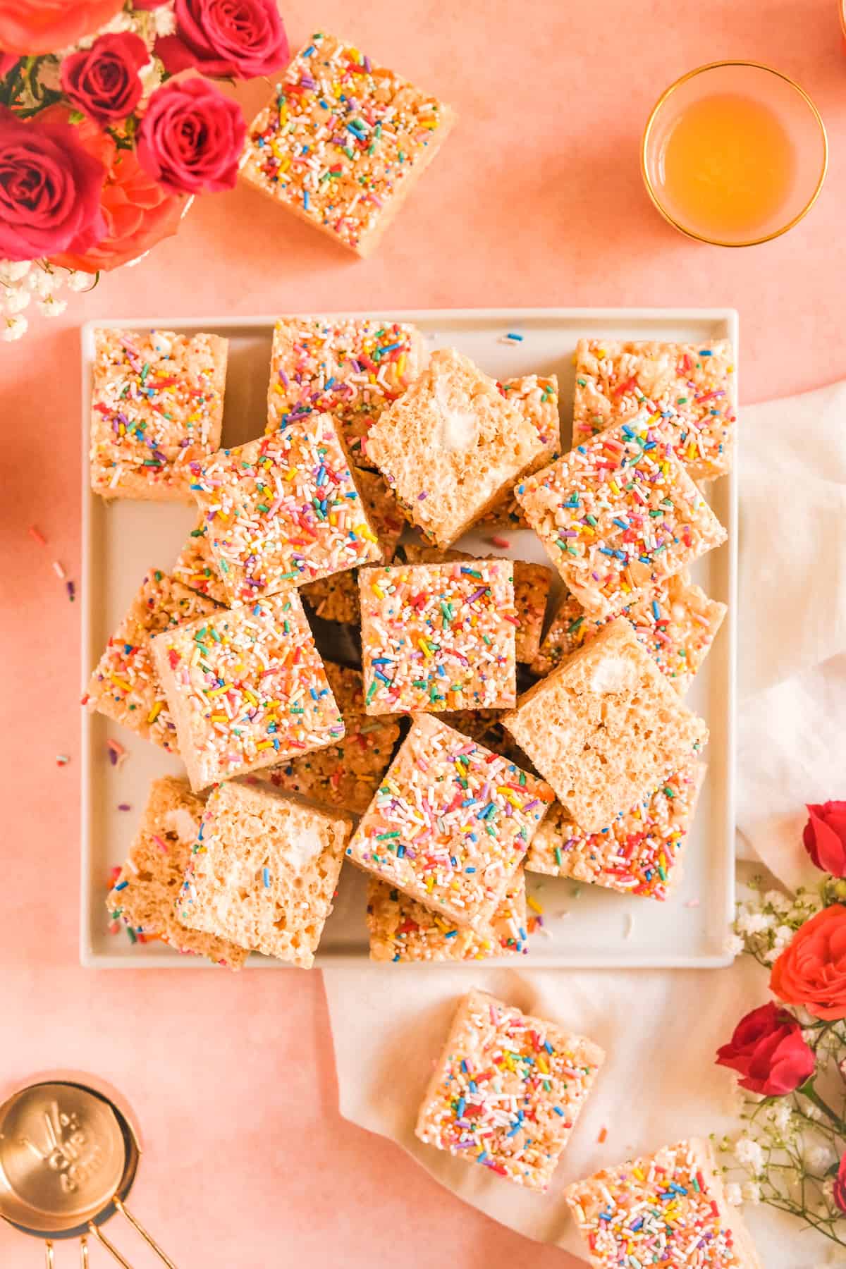 Brown Butter Rice Krispie Treats stacked on a plate with a glass of orange juice, measuring bags, and flowers on the table