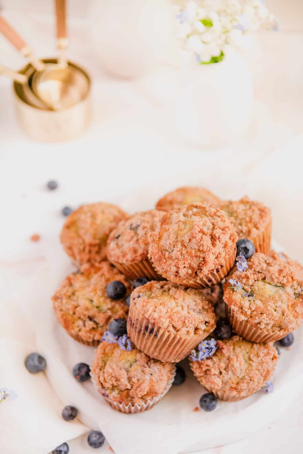 blueberry muffins with cinnamon streusel stacked up on a plate with forget me nots and blueberries on the plate
