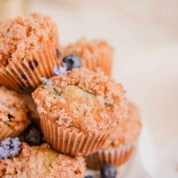 blueberry muffins with cinnamon streusel stacked up on a plate with forget me nots and blueberries on the plate