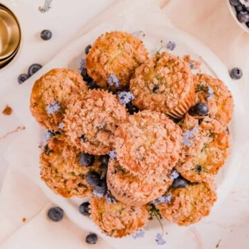 blueberry muffins with cinnamon streusel stacked up on a plate with forget me nots and blueberries on the plate