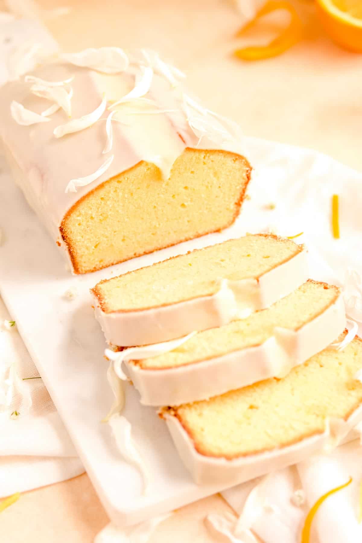 a partially sliced, glazed citrus cream cheese pound cake from a 45 degree angle and oranges in the background.