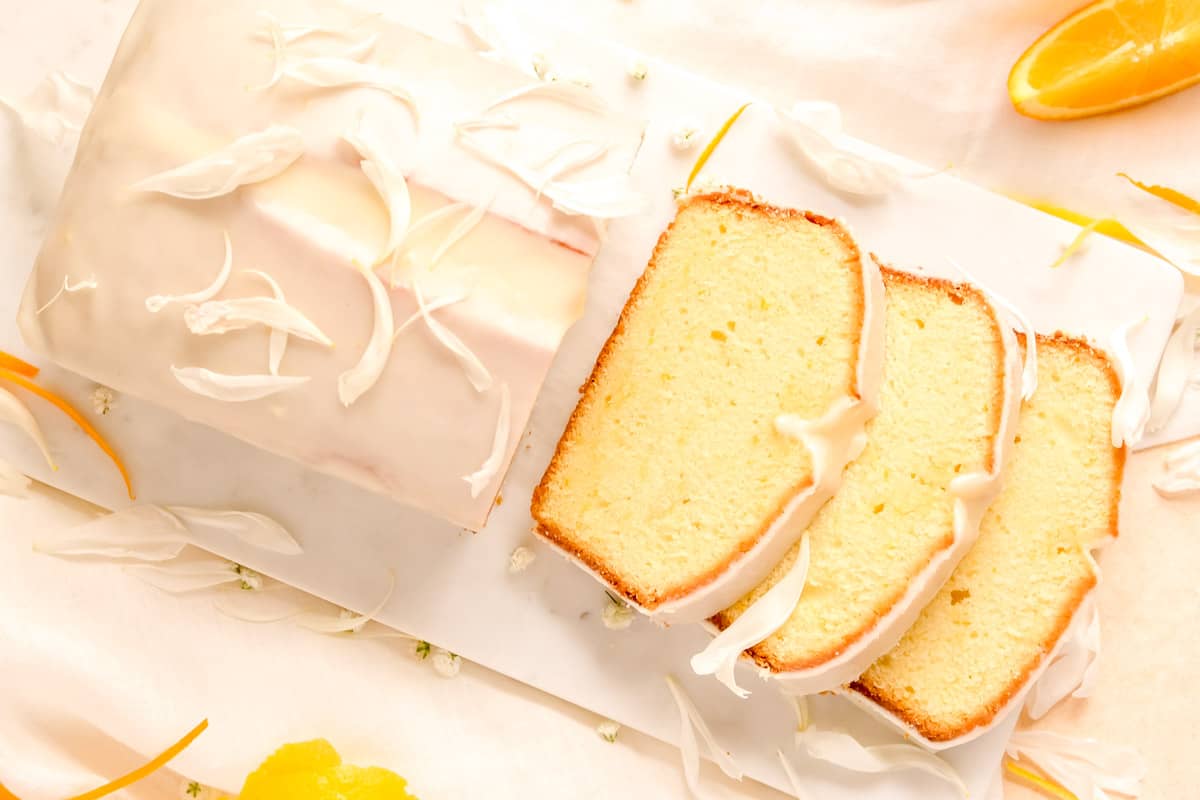 a partially sliced citrus cream cheese pound cake from above on a marble board with orange and lemon slices on the table.