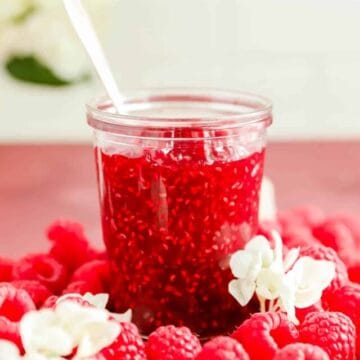 a clear glass jar of french raspberry jam surrounded by raspberries.