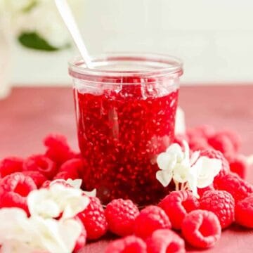 a clear glass jar of french raspberry jam surrounded by raspberries.