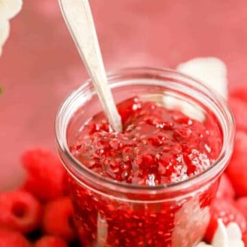a clear glass jar of homemade raspberry jam surrounded by raspberries and flowers.