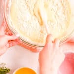a hand holding a spatula folding flour into a fluffy batter with a bowl of melted butter on the side.