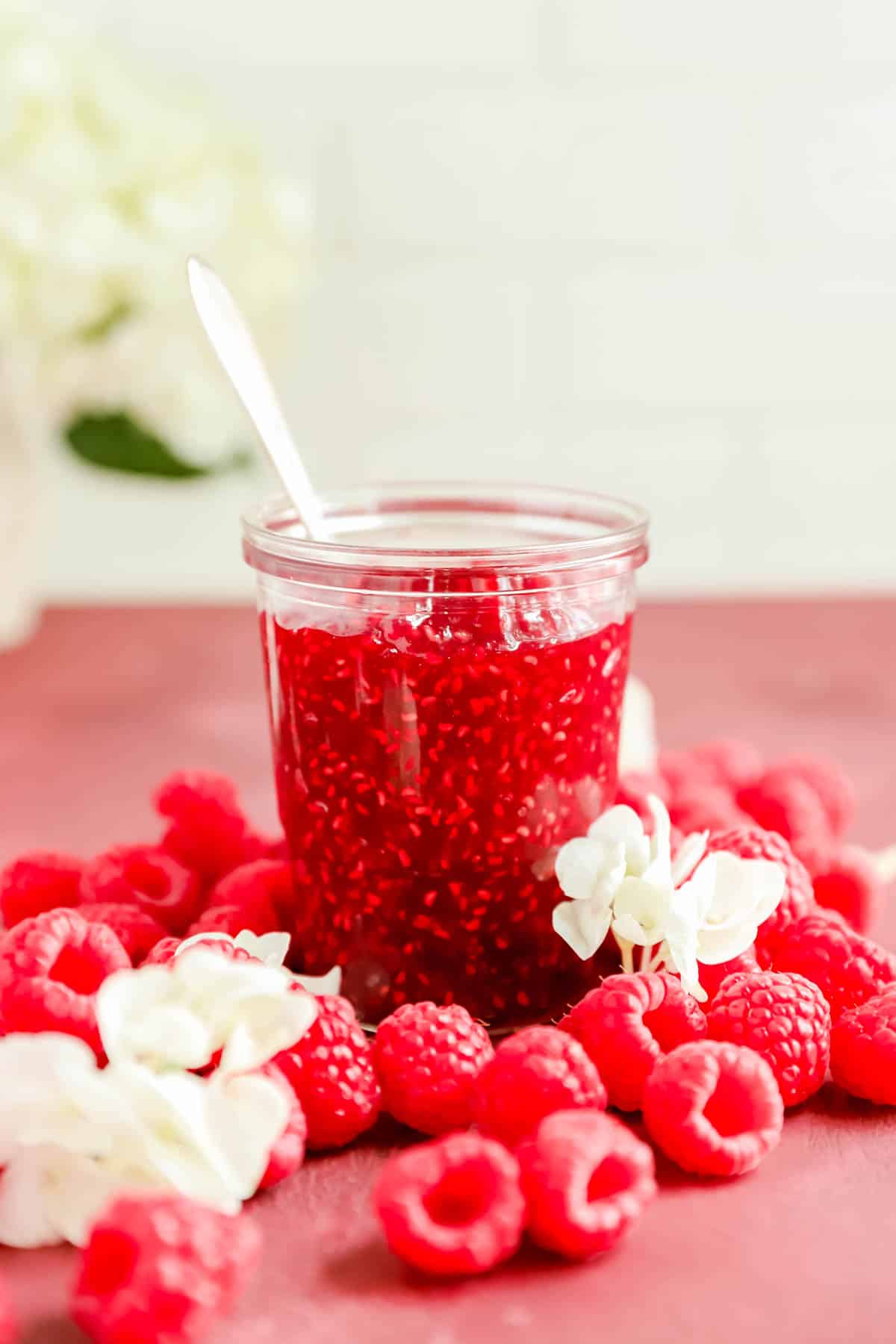 a clear glass jar of french raspberry jam surrounded by raspberries.