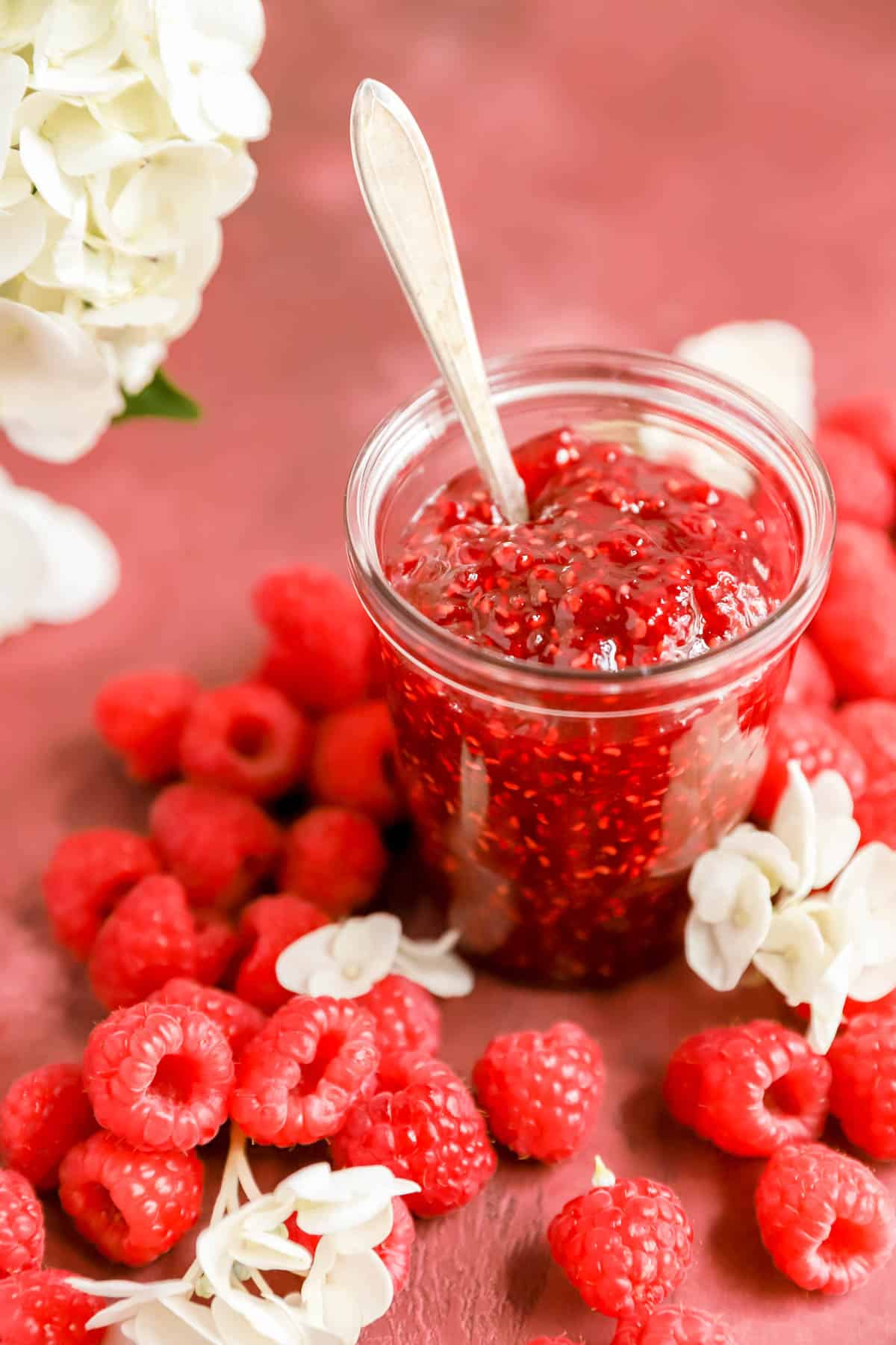 a clear glass jar of homemade raspberry jam surrounded by raspberries and flowers.