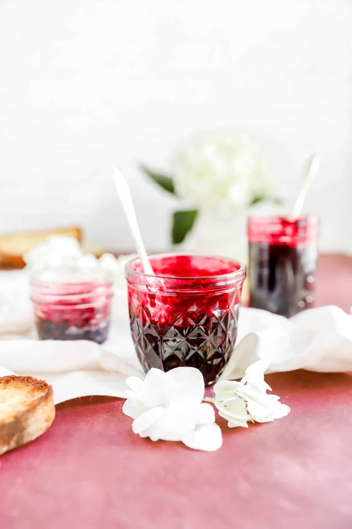 3 jars of black raspberry jam with toast on a burgundy table with flowers scattered around.