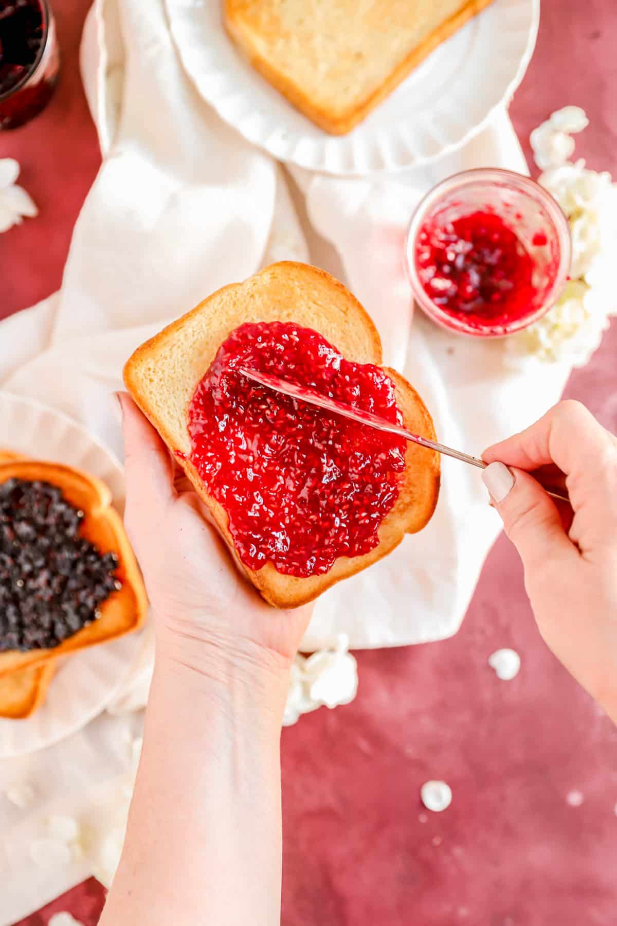 a hand holding a piece of toast and spreading homemade red raspberry jam on it.