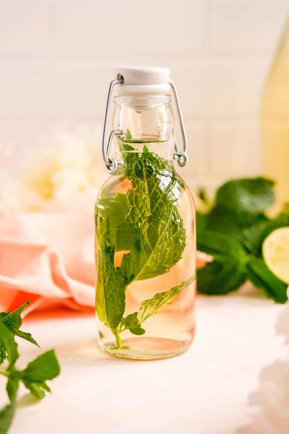 a bottle of mint simple syrup with a mint leaf sprig in it and flowers in the background.