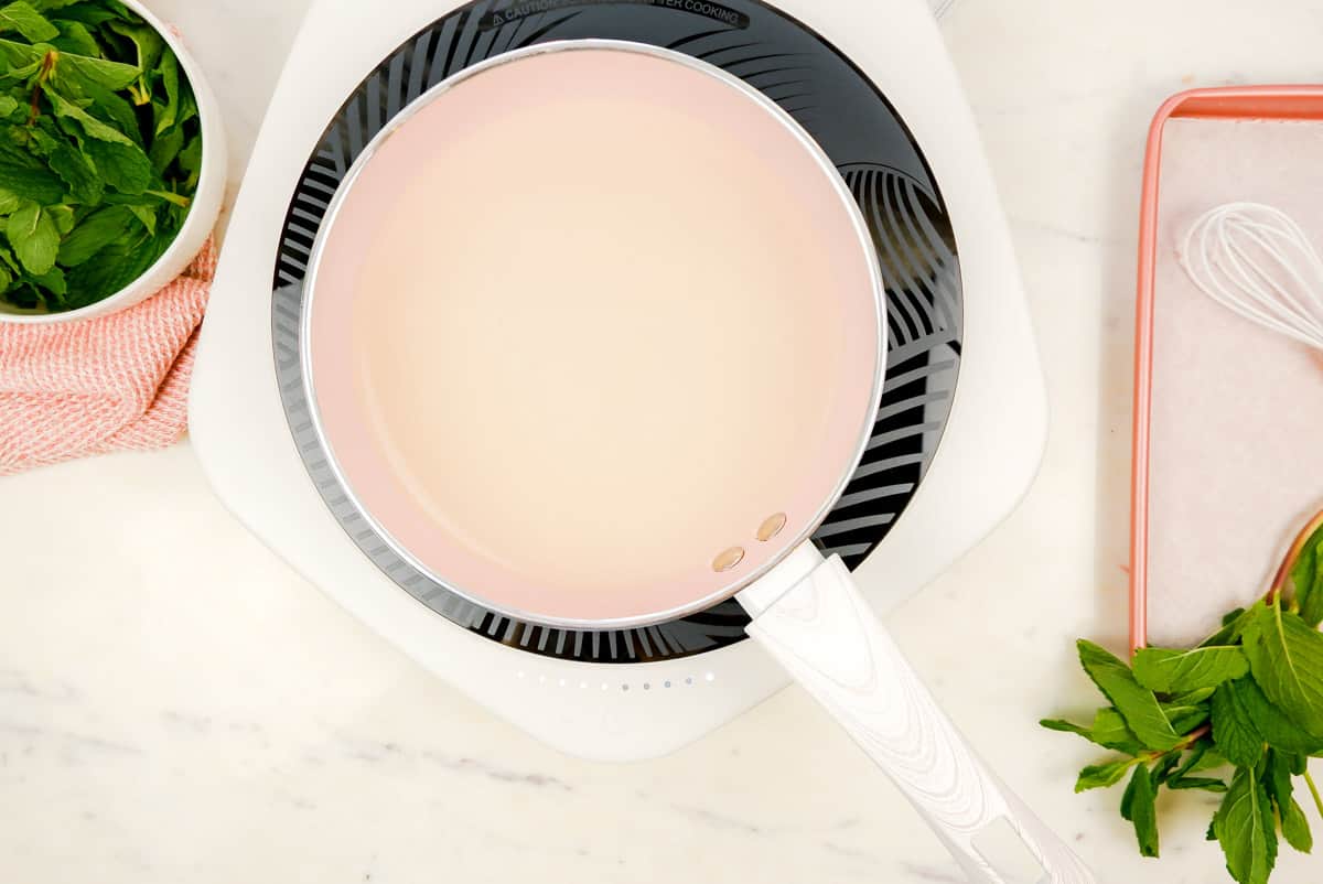 a pan on an induction stove with water and sugar in it with mint leaves in a bowl on the side.