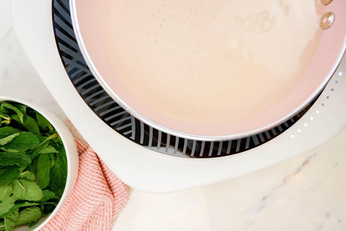 a pink pan on an induction stove with water and sugar in it starting to bubble with mint leaves in a bowl on the side.