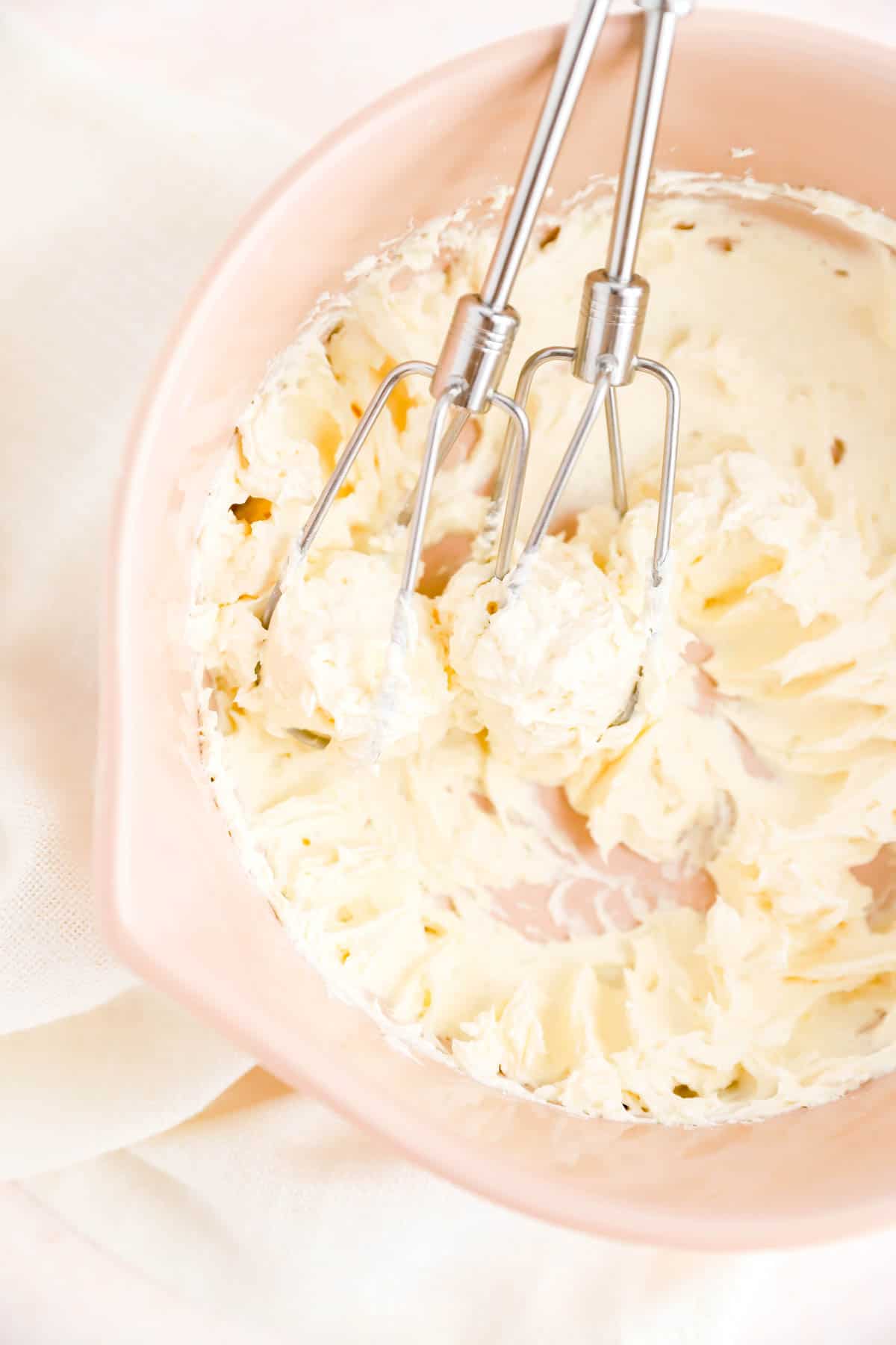 a tan bowl of whipped butter and cream cheese with silver mixer beaters in it.