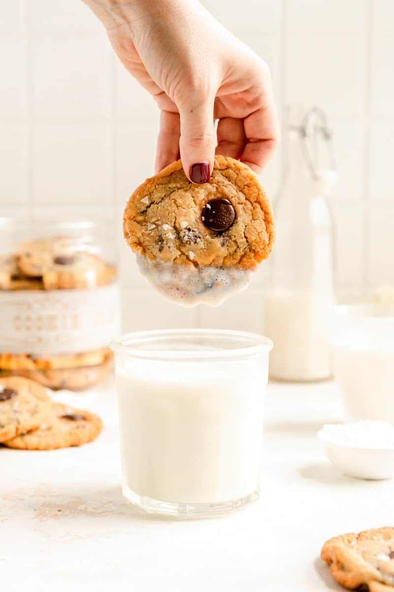 a hand dunking a cookie into a cup of milk with a cookie jar and milk jug.