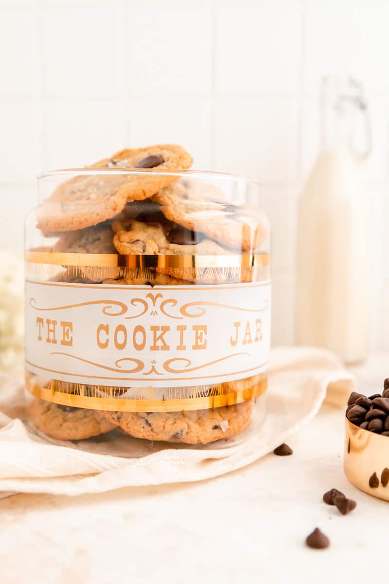 a vintage clear, gold, and white cookie jar filled with chocolate chip cookies.
