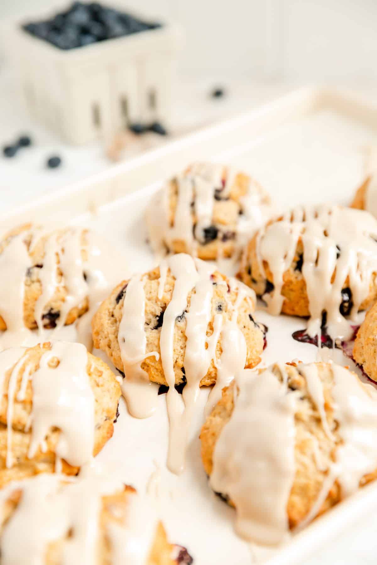 a white baking sheet filled with glazed baked scones with blueberries in the background