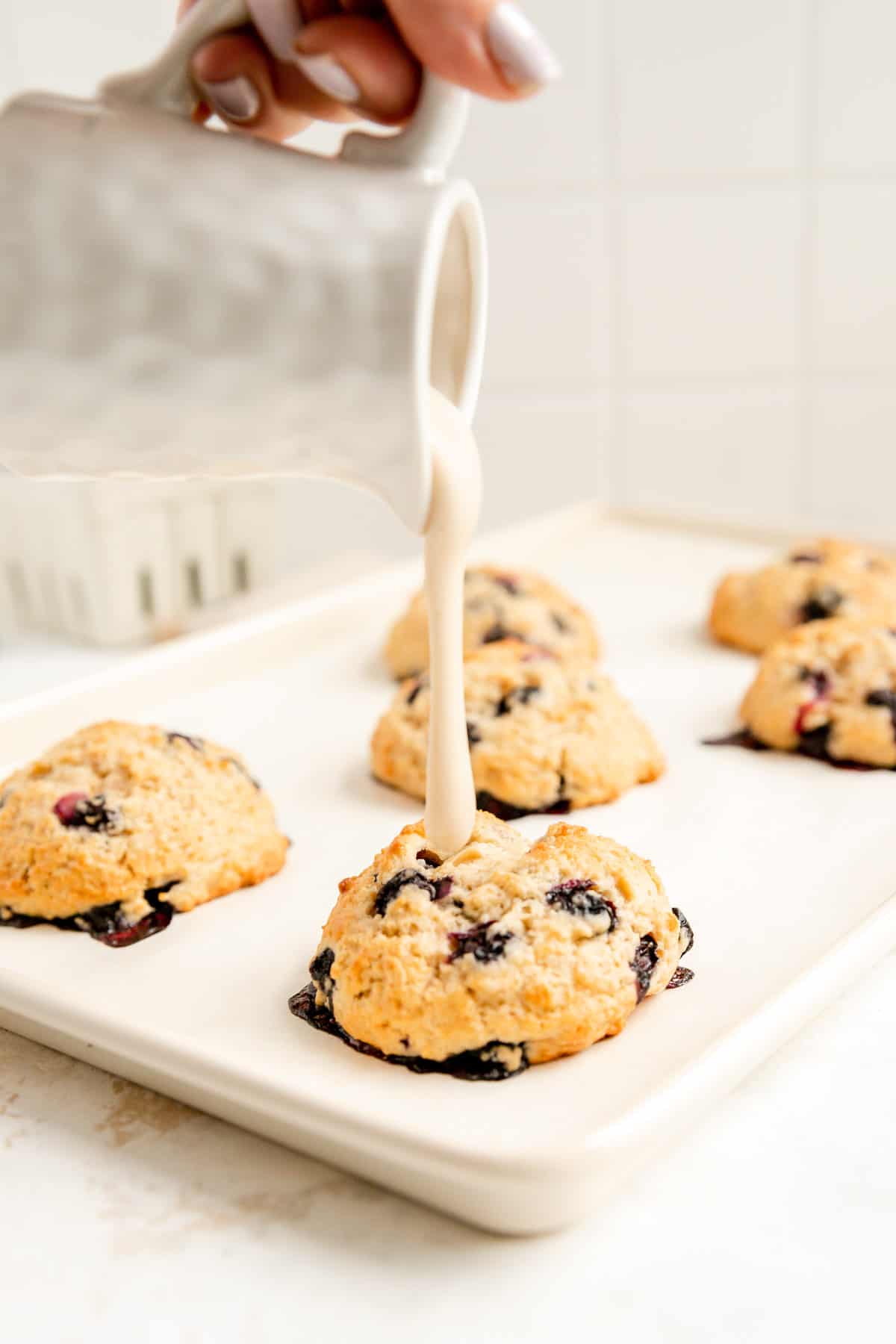 a hand pouring maple glaze out of a white pitcher onto a tray of baked scones