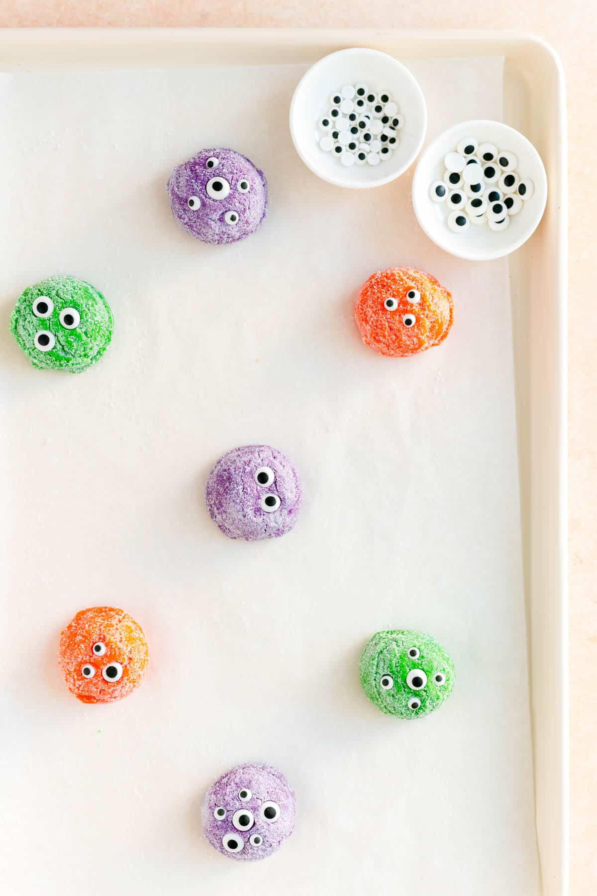 purple green and orange dough balls with candy eyes on top on a white baking sheet