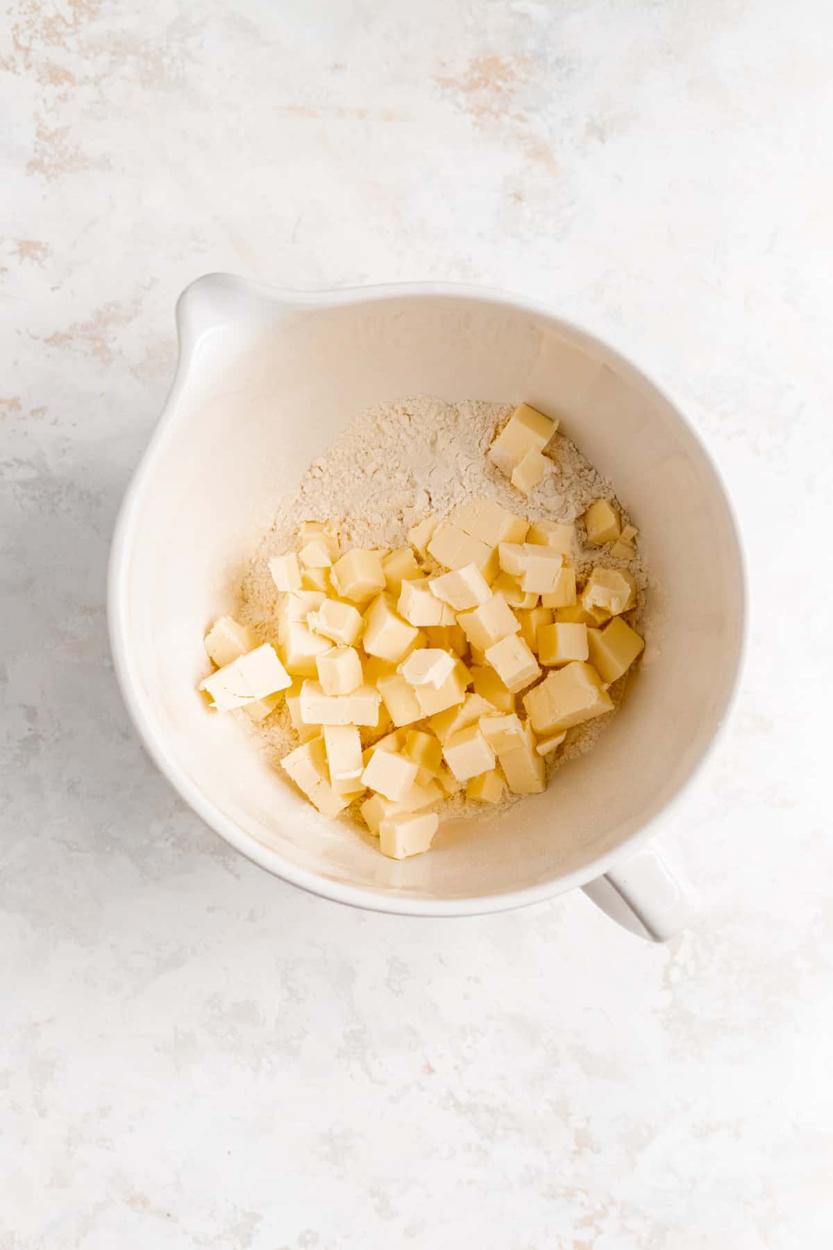 cold cubed butter on a pile of flour in a white mixing bowl on a white plaster background