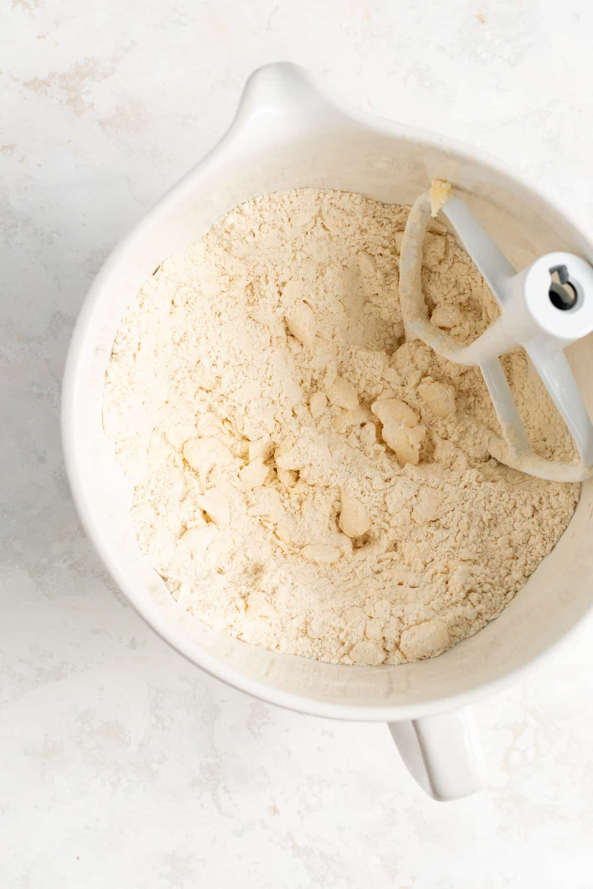 cold butter flakes mixed into flour in a white mixing bowl with a white mixer beater