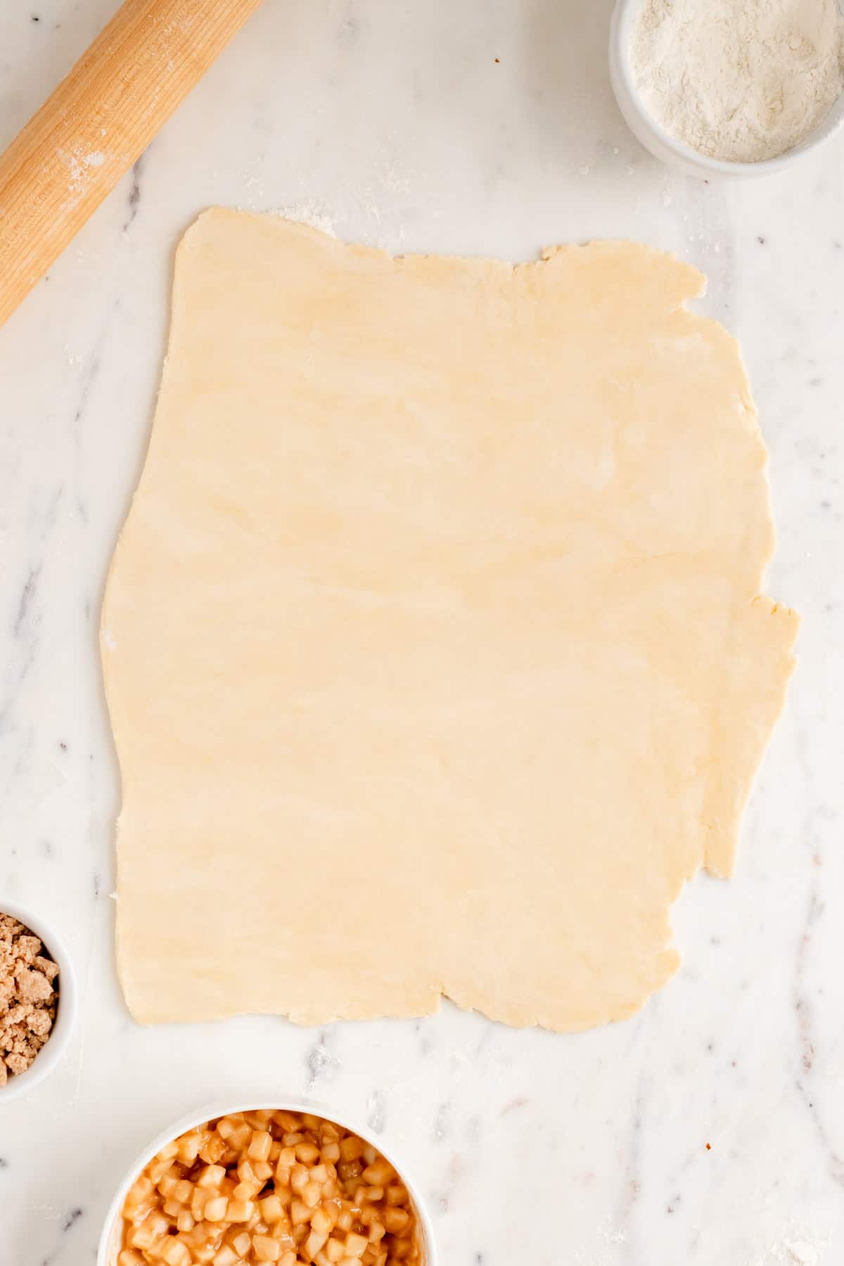 pie dough rolled out on a white marble table with apple filling, streusel and flour in bowls