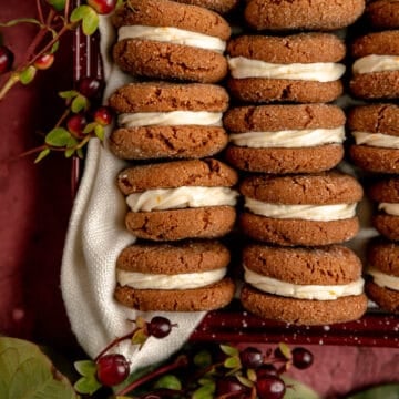 Lots of ginger creme sandwich cookies in a red pan with red berries and leaves around them