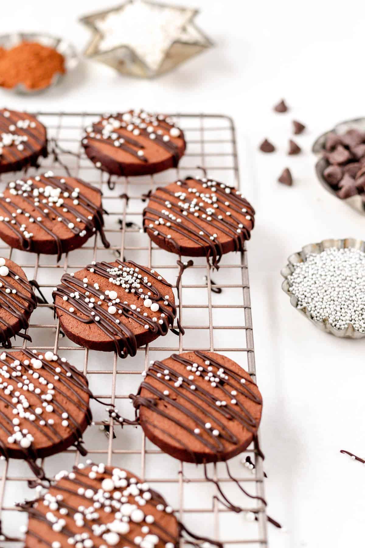 Decorated chocolate butter cookies on a colling rack with tins of sprinkles, chocolate chips, and cocoa