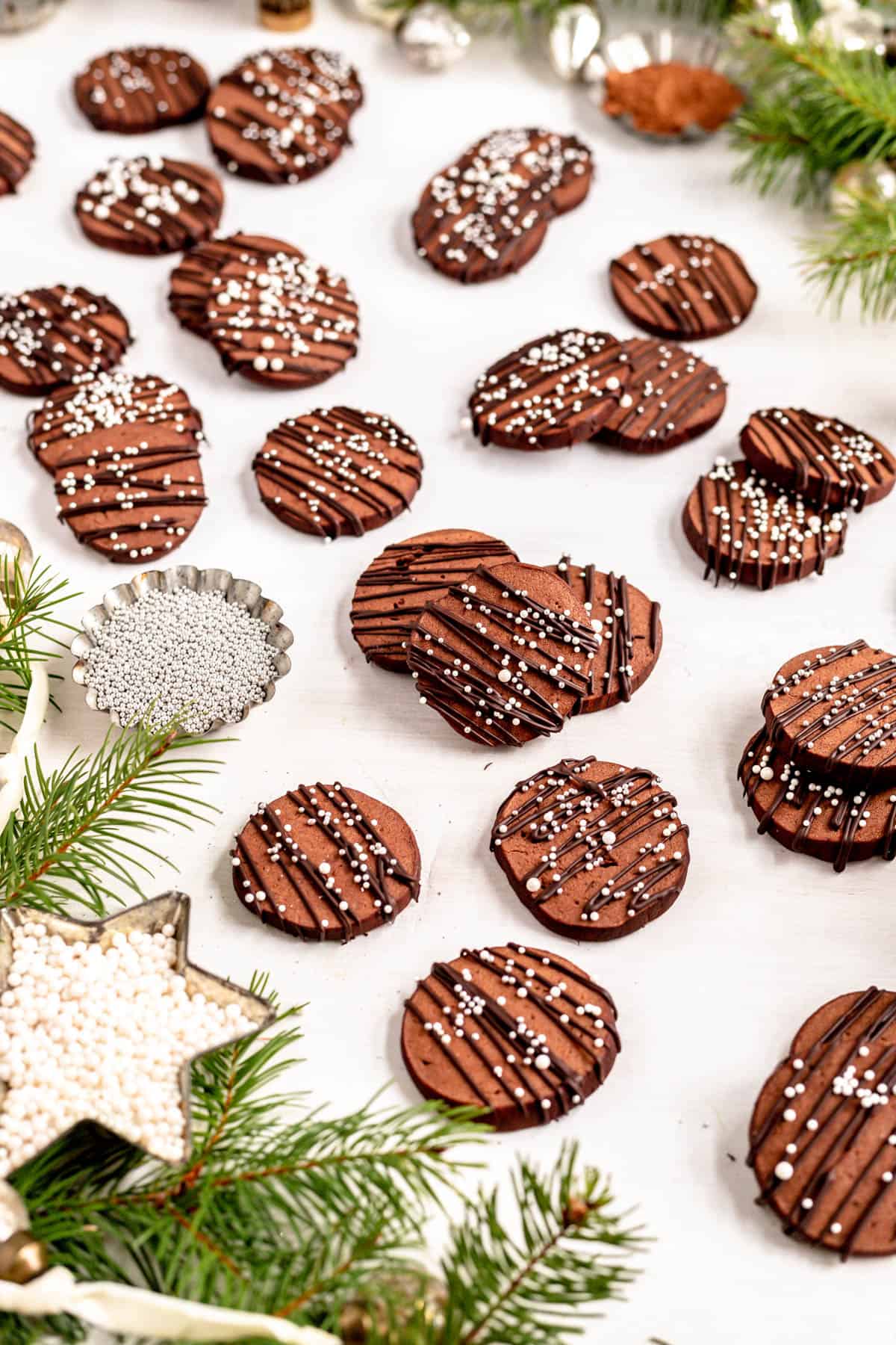 chocolate butter cookies with chocolate drizzle and white sprinkles scatter on a table with evergreen branches.