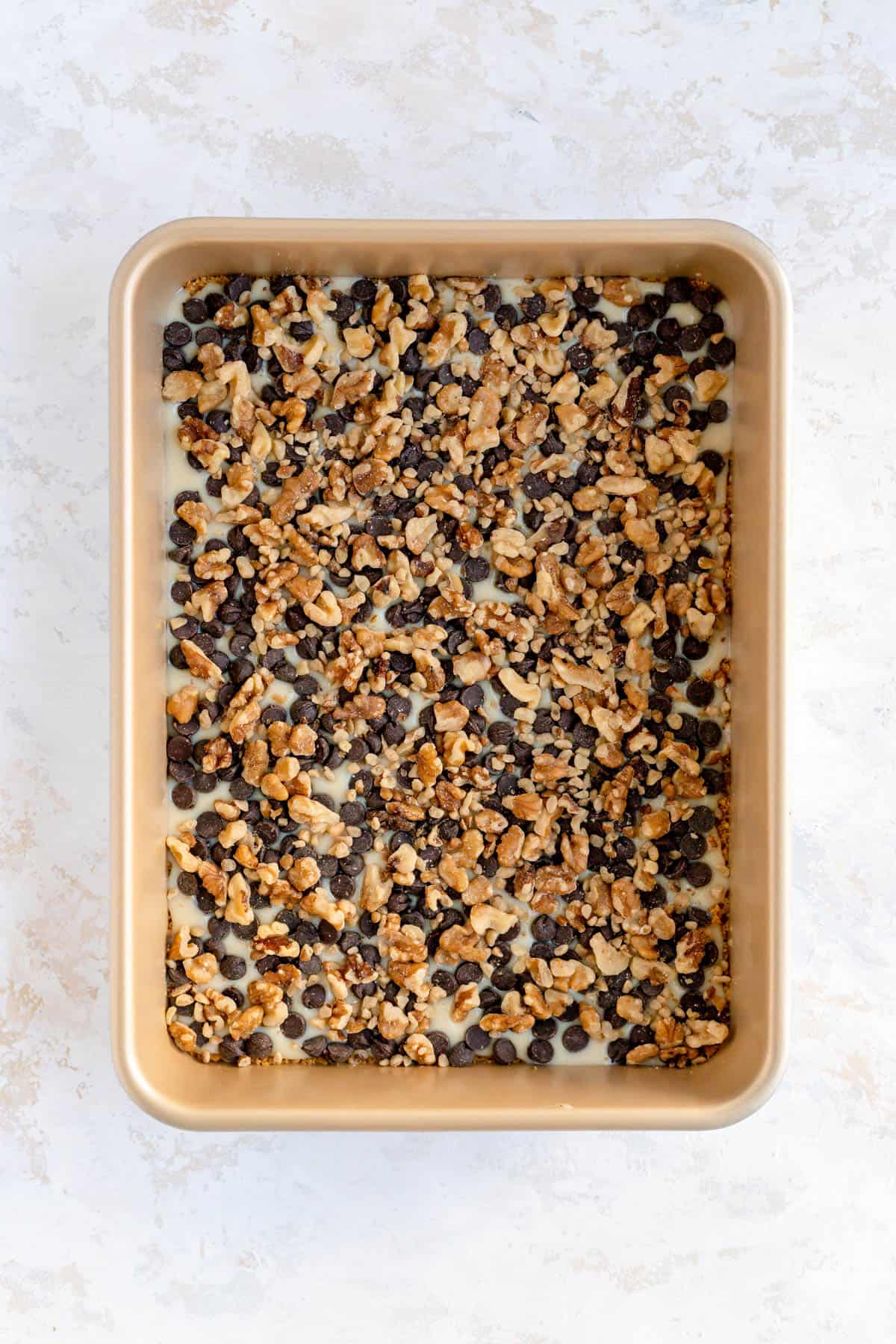 Walnuts sprinkled over chocolate chips and condensed milk in a gold 9 x 13 pan