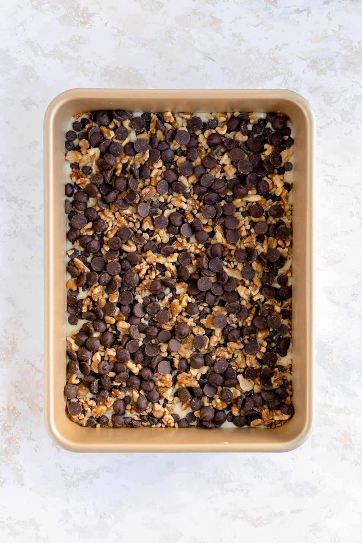 Chocolate chips sprinkled over walnuts and condensed milk in a cold 9 x 13 pan