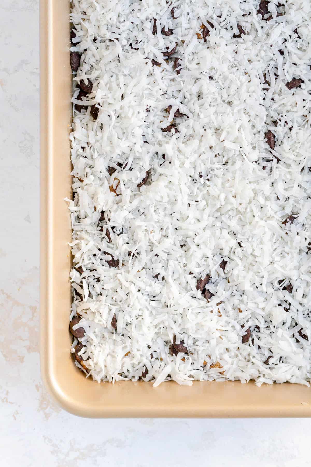 Coconut sprinkled over chocolate chips in a gold pan of seven layer bars