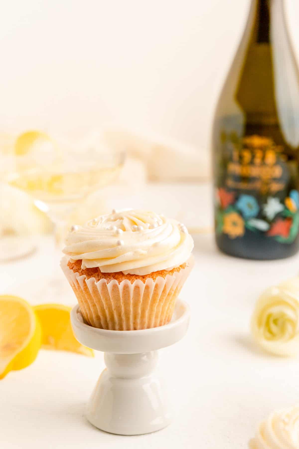 A single cupcake on small stand with lemon flowers and a green prosecco bottle behind it.