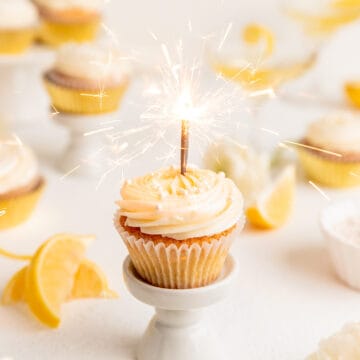 A single cupcake on a small stand with a lit sparkler candle in it sparkling.