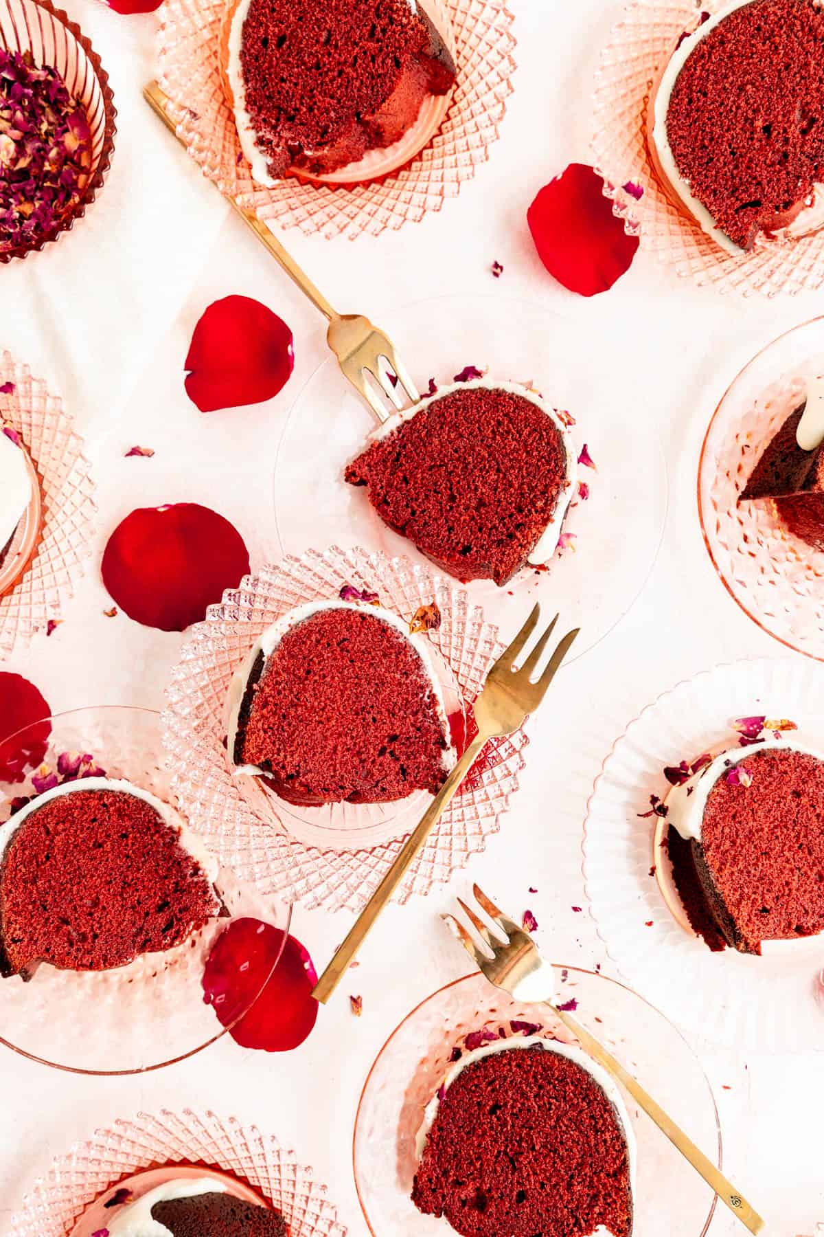 Slices of red velvet Bundt cake on pink crystal plates with gold forks and rose petals.