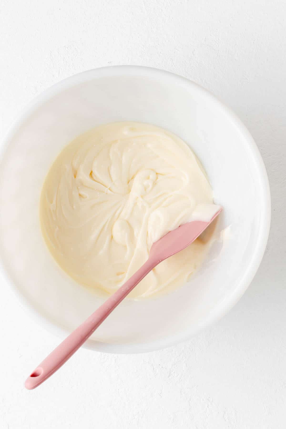 Cream cheese glaze in a white bowl with pink spatula on white background.