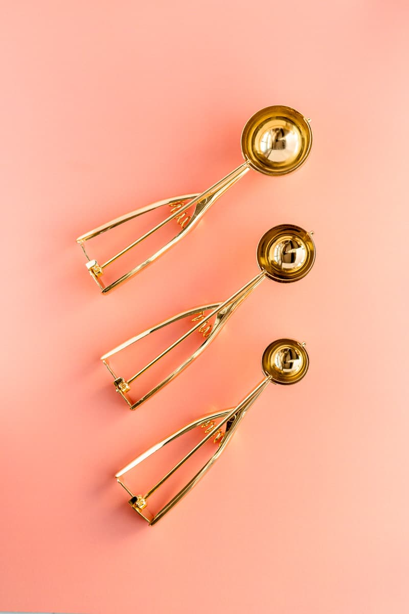 Three sizes of gold ice cream scoops on coral background.