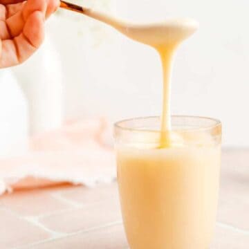 Hand holding spoon with white chocolate sauce dripping off into clear glass container.