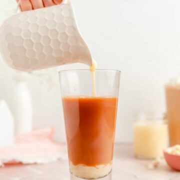 Hand holding white honeycomb pitcher pouring white chocolate sauce in tall glass of coffee.