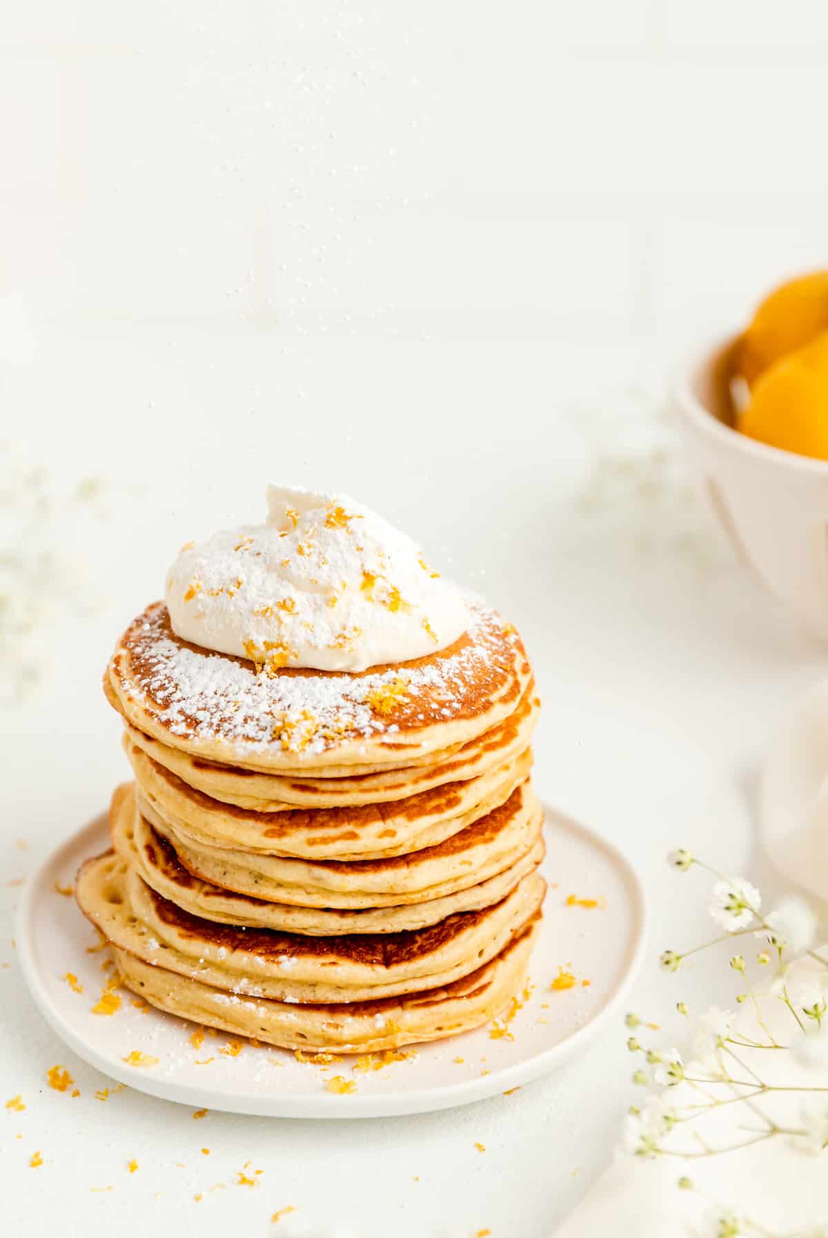 lemon pancakes stacked on white plate topped with whipped cream being sprinkled with powdered sugar.