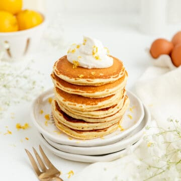 45° shot of lemon pancakes stacked up with whip cream and lemons in bowl in back.